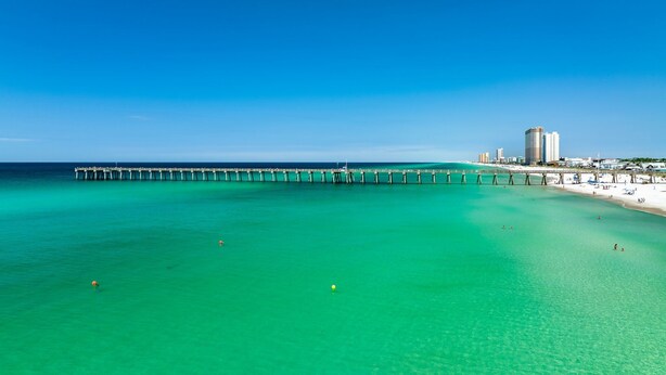 A pier extending into turquoise waters with a beach and buildings in the background.