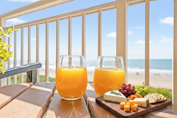 A table with two glasses of orange juice, a cheese board with various cheeses and crackers, and a view of the beach and ocean.