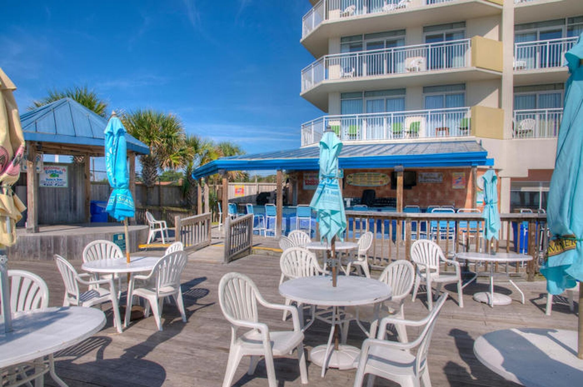 Outdoor seating with white plastic chairs and tables, a pool area with blue umbrellas, and a building with balconies.