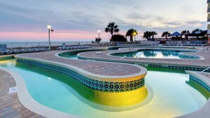 A swimming pool with a tiled edge and a curved design, surrounded by a deck area with lounge chairs and a view of the ocean at sunset.