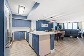 A modern kitchen with blue cabinets, a central island, and a dining area with a sofa and table. - Ocean Bay Club 1109-Pool, Hot Tub, Lazy River-4BD (North Myrtle Beach)