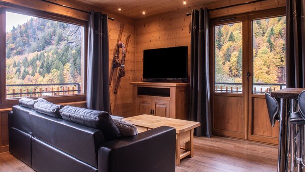 Ein gemütliches Blockhaus-Interieur mit Holzbalkendecke, einem großen Fenster mit Blick auf einen Wald, einer Ledersofa, einem Holztisch und einem Fernseher.