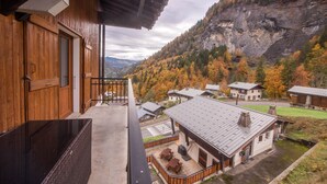 Ein Holzhaus mit Balkon, das auf ein Tal mit herbstlich gefärbten Bäumen und einem felsigen Berg blickt.