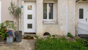 Ein zweistöckiges Gebäude mit weißer Fassade, einem kleinen Fenster im Erdgeschoss und einem größeren Fenster im Obergeschoss. Im Erdgeschoss befindet sich eine Tür mit kleinem Fenster und ein Briefkasten.