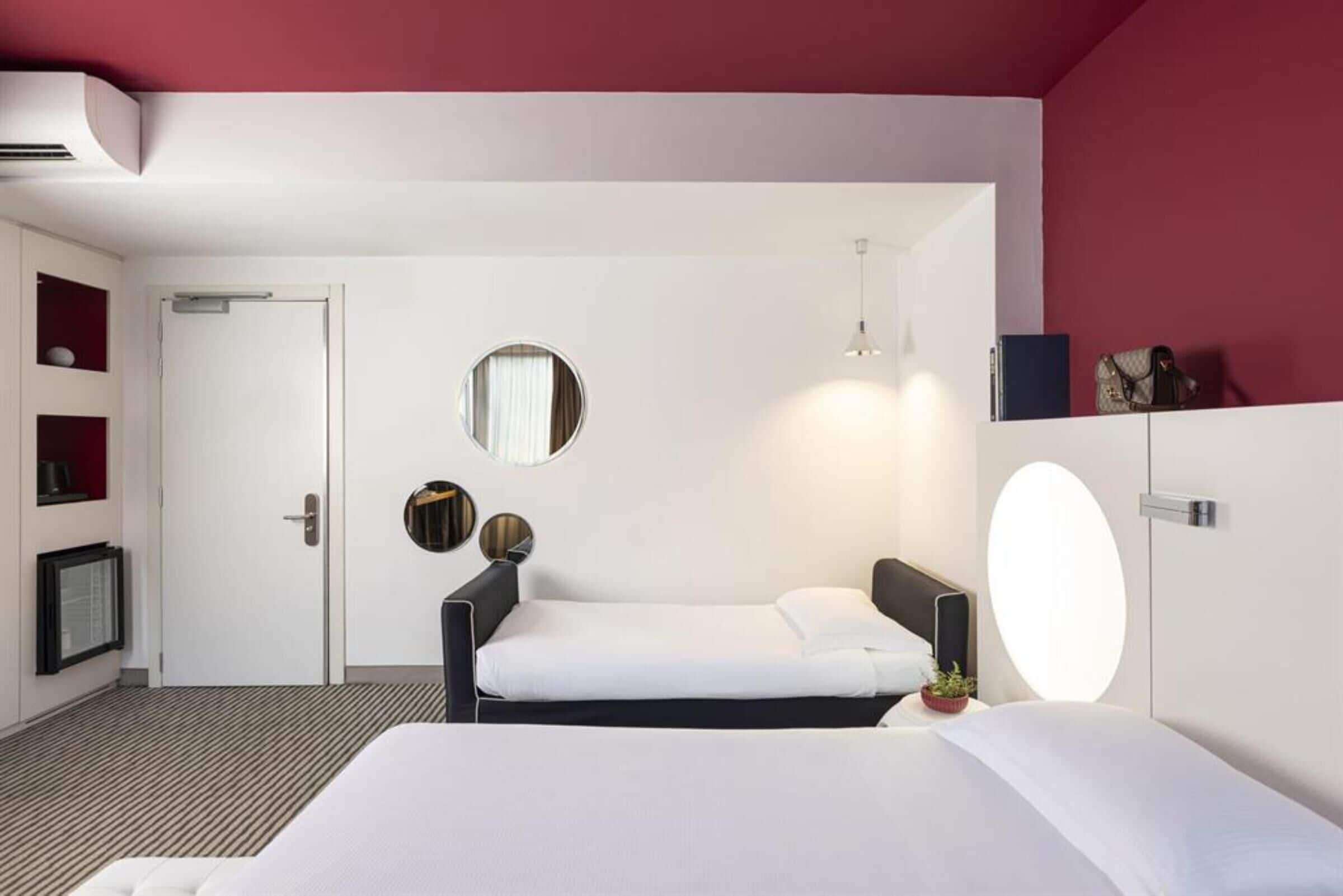 A hotel room with a large bed, a smaller bed, a mirror, a shelf with items, and a door.