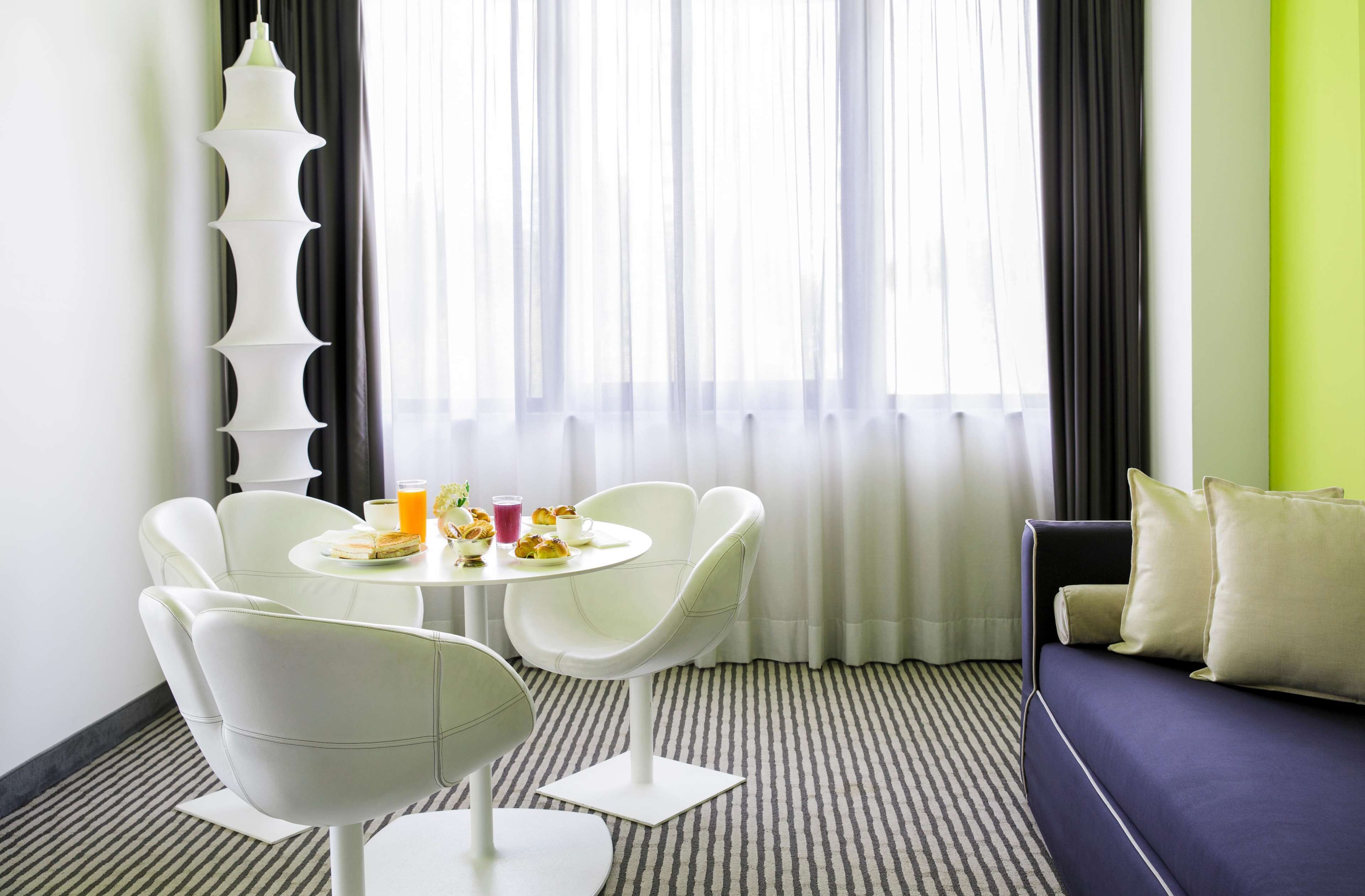 Ett modernt hotellrum med en matplats som inkluderar ett runt bord och vita stolar, en soffa och ett stort fönster med tunna, genomskinliga gardiner.