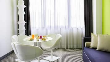 Une chambre d’hôtel moderne dotée d’un coin repas comprenant une table ronde et des chaises blanches, un canapé et une grande fenêtre avec des rideaux légers.