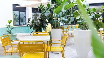 An outdoor seating area with yellow chairs and white tables, surrounded by large potted plants.