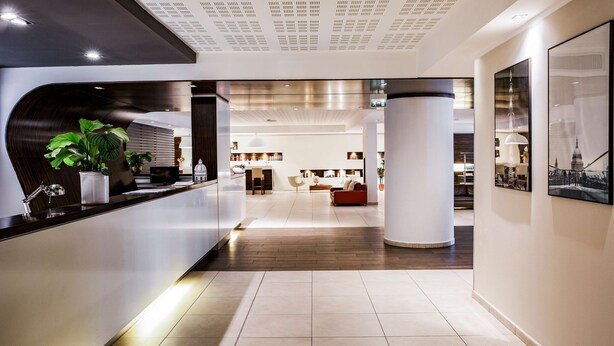Een moderne hotellobby met een receptiebalie, grote kamerplanten, een zithoek met een rode bank en een uitzicht op het interieur door een glazen wand.