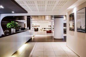 Een moderne hotellobby met een receptiebalie, grote kamerplanten, een zithoek met een rode bank en een uitzicht op het interieur door een glazen wand.