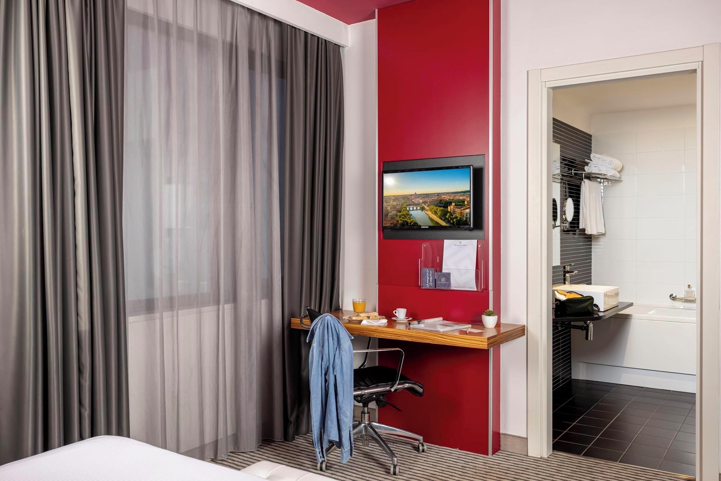 A hotel room with a bed, a desk with a chair, a TV mounted on the wall, and a bathroom visible through an open door.