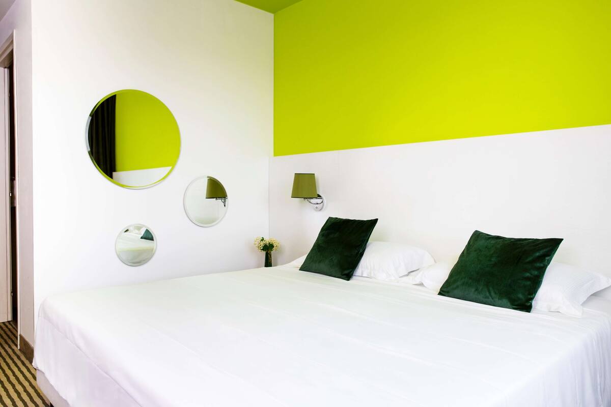 Une chambre d’hôtel avec un lit, un miroir rond, deux lampes fixées au mur et un oreiller vert.