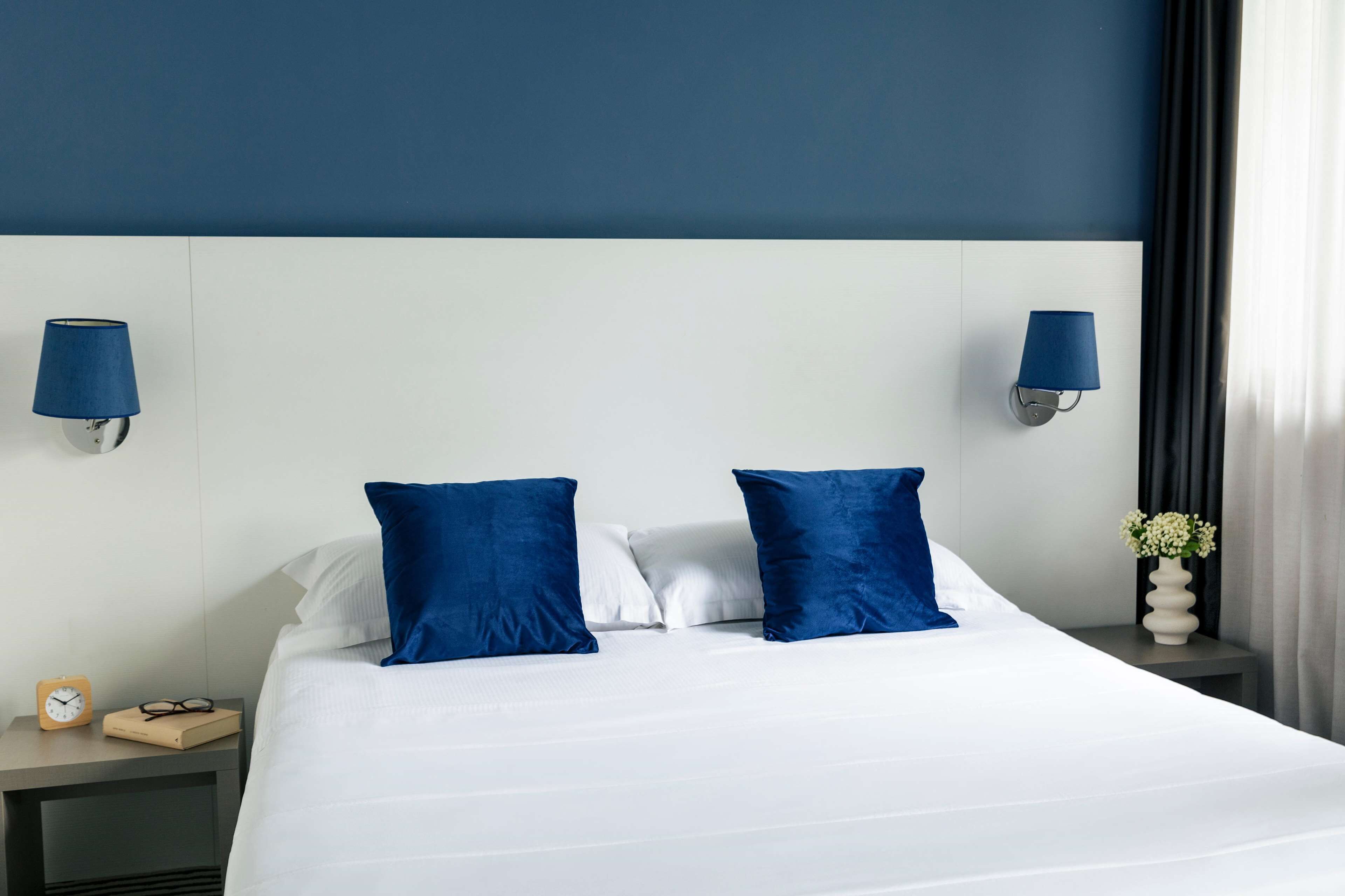 Habitación de hotel con una cama grande, dos almohadas azules, un cabecero blanco, una mesita de noche con una lámpara, un reloj y un jarrón con flores.