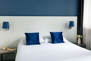 Et hotelværelse med en stor seng, to blå puder, en hvid sengegavl, et natbord med en lampe, et ur og en vase med blomster.