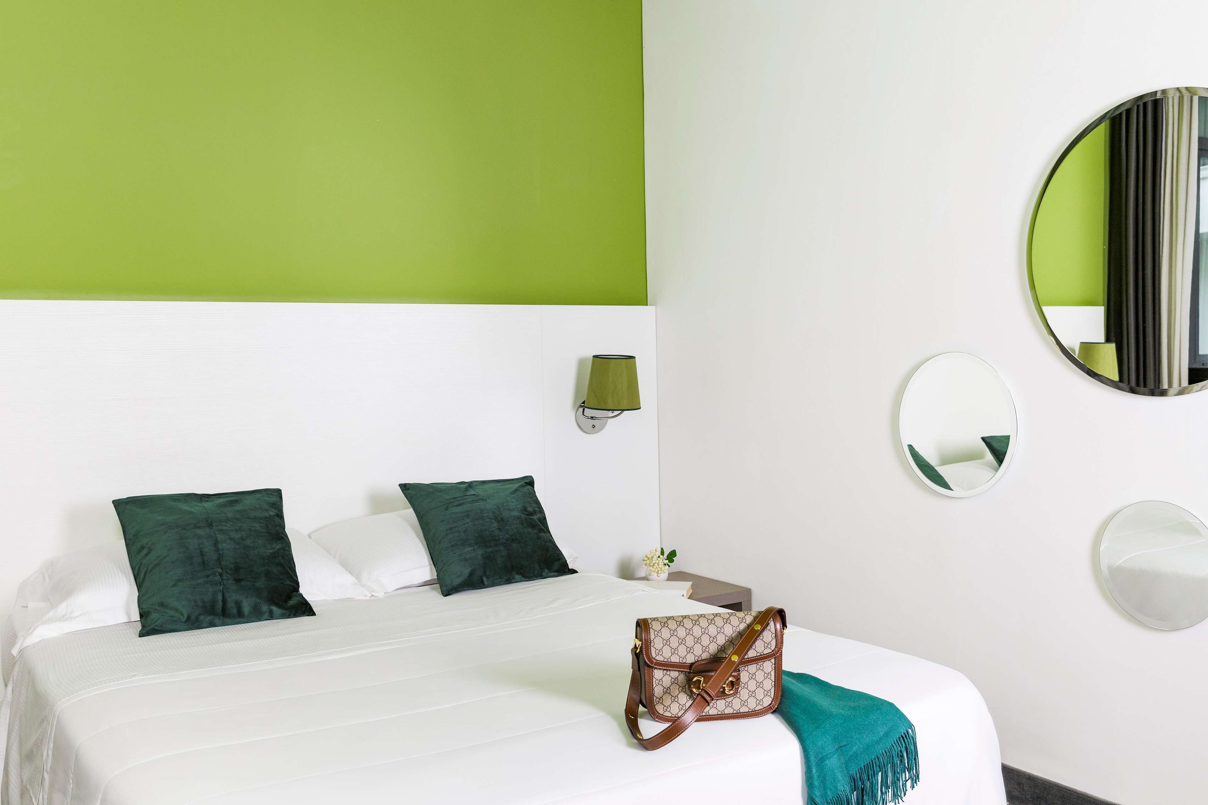 Une chambre d’hôtel avec un lit, des oreillers verts, un plaid vert, un sac à main, une table de chevet, une lampe et deux miroirs muraux.