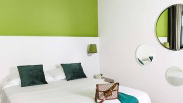 Une chambre d’hôtel avec un lit, des oreillers verts, un plaid vert, un sac à main, une table de chevet, une lampe et deux miroirs muraux.