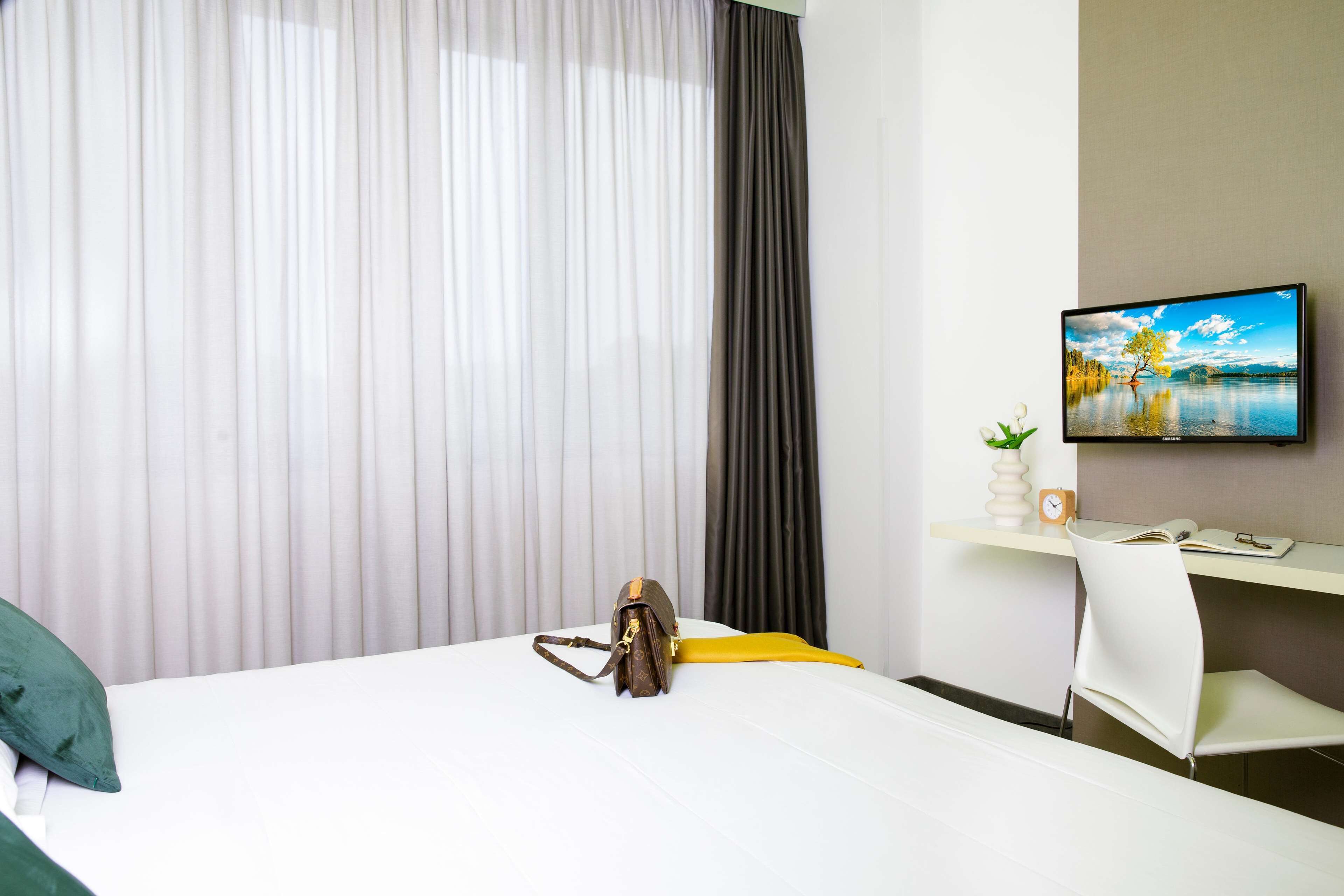 A hotel room with a bed, a desk with a computer, a TV on the wall, and a window with curtains.