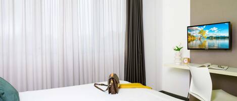 Habitación de hotel con cama, escritorio con ordenador, televisión en la pared y ventana con cortinas.