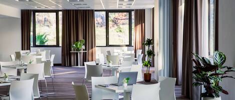 Una sala de conferencias moderna con sillas y mesas blancas, ventanas grandes con cortinas y plantas en macetas.