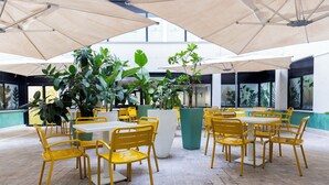 Een binnenplaats met gele stoelen en tafels, grote potplanten en een betegelde vloer.