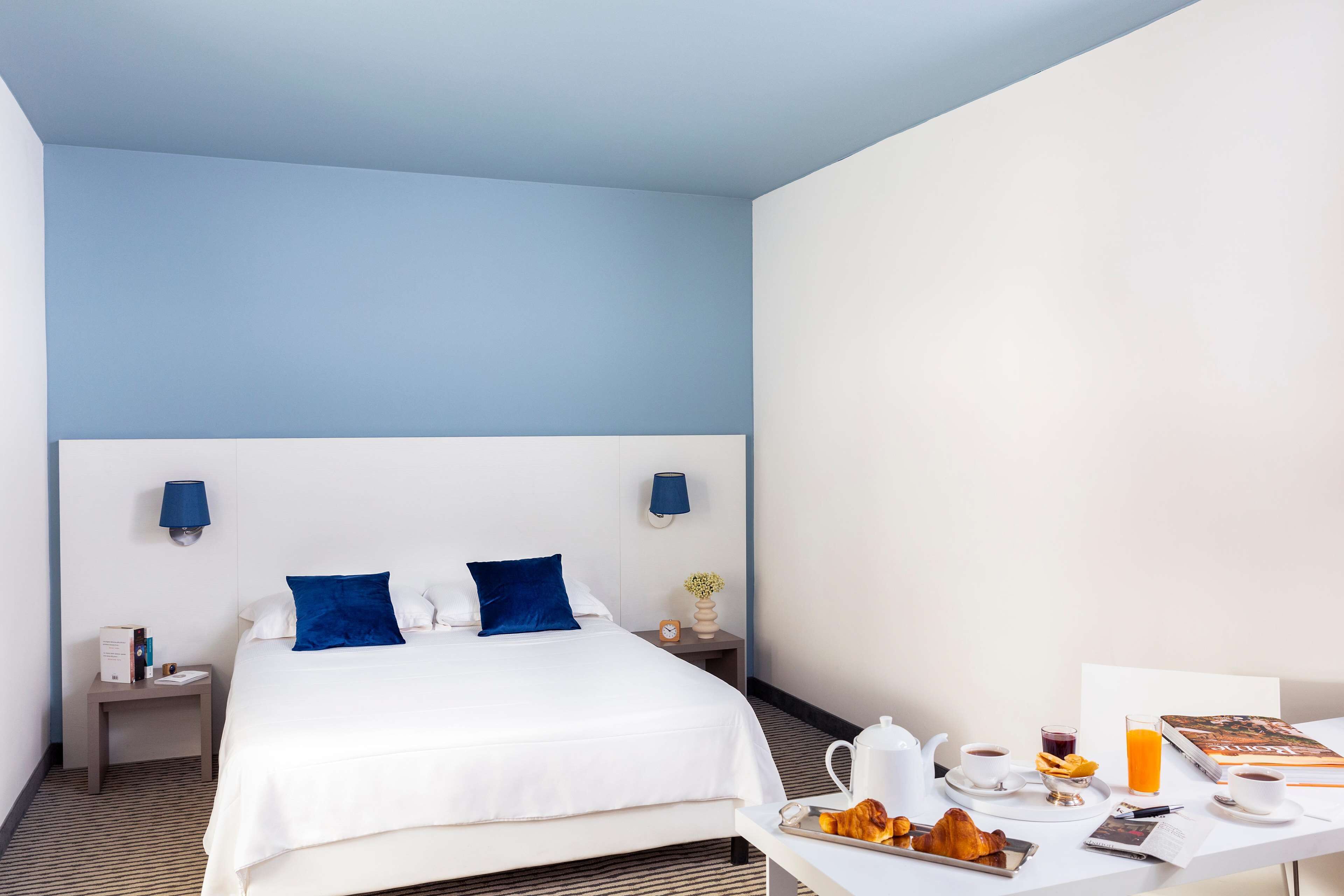 A hotel room with a bed, bedside tables, a breakfast table with croissants, coffee, and juice.