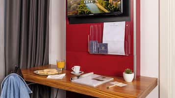 Quarto de hotel com escrivaninha de madeira, televisão na parede, parede vermelha, janela com cortinas, uma planta e um tabuleiro de café da manhã com sanduíche, suco e uma xícara.