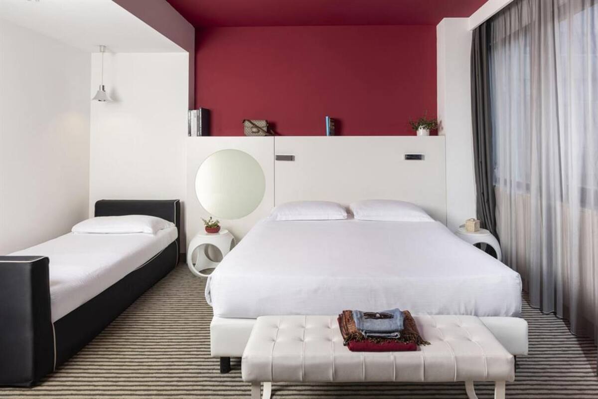 Une chambre d’hôtel avec un grand lit, un lit plus petit, une table de chevet avec une lampe et une fenêtre avec des rideaux.