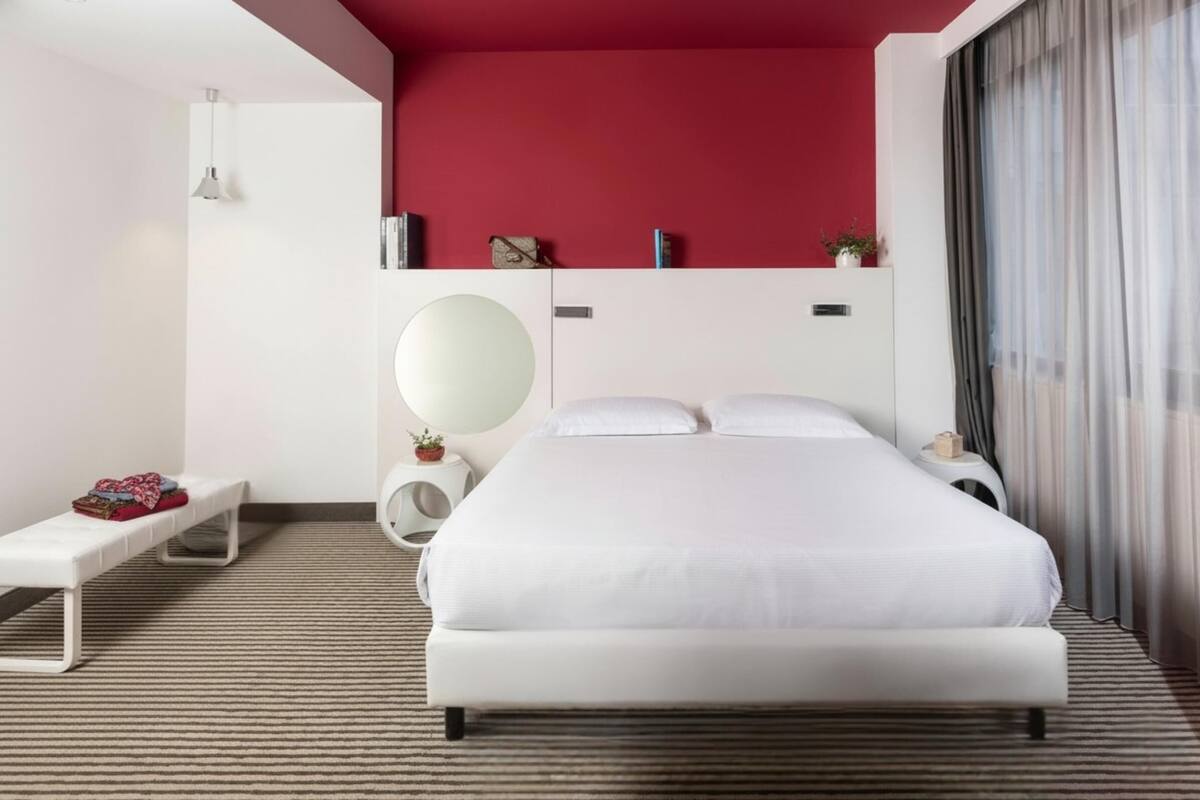 Une chambre d’hôtel moderne avec un grand lit, un banc, un miroir rond et une table de chevet avec une lampe.