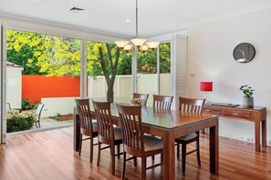 Un coin repas avec une table et des chaises en bois, un luminaire suspendu, une horloge au mur et une vue sur un jardin aux feuillages jaunes.