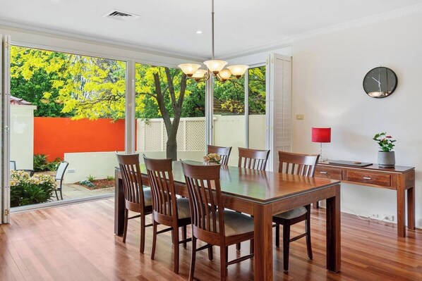 Uma sala de jantar com mesa e cadeiras de madeira, uma luminária pendente, um relógio na parede e vista para um jardim com folhagem amarela.