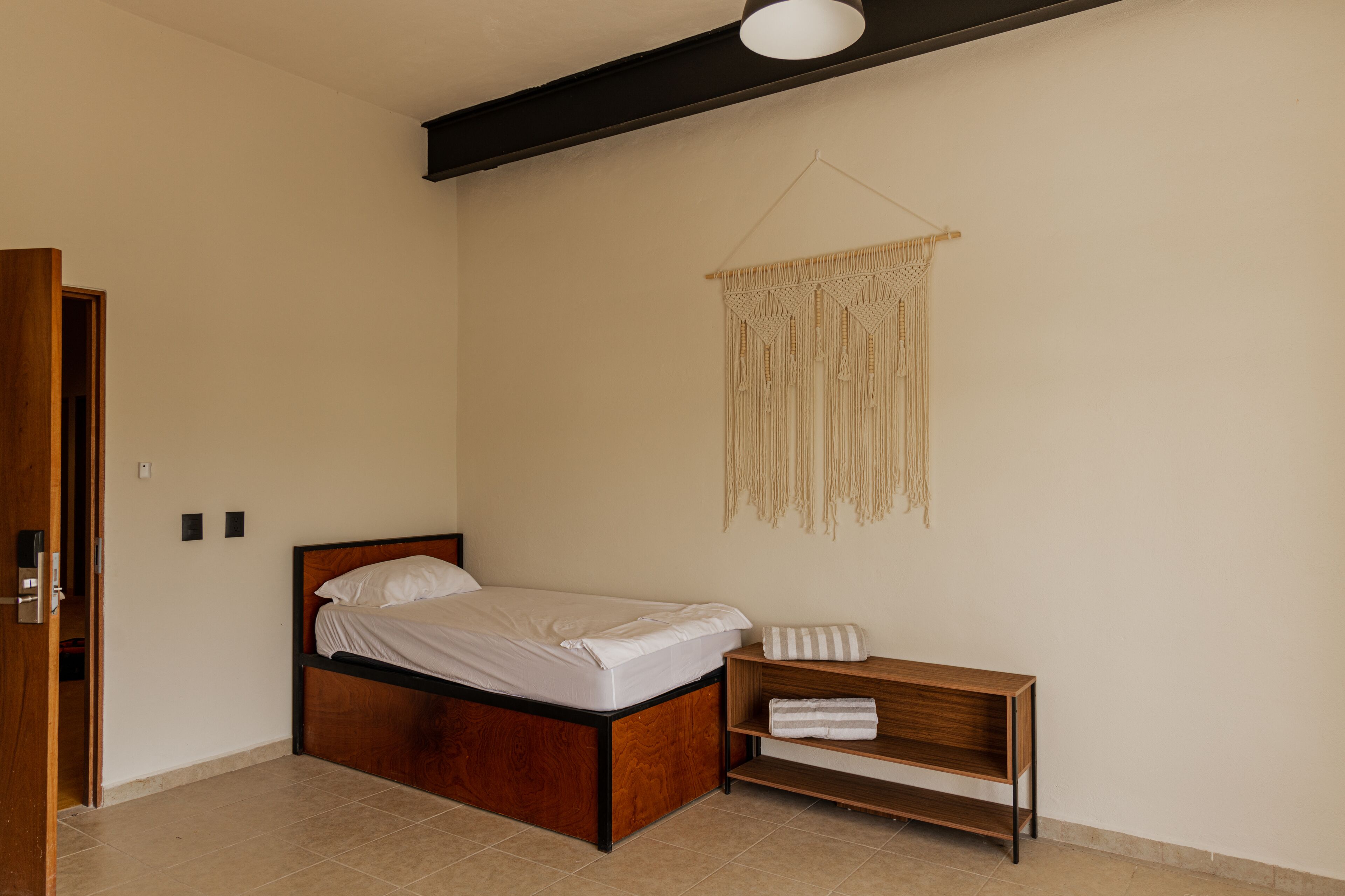A bedroom with a wooden bed, a small wooden nightstand, a macrame wall hanging, and a door to the left.