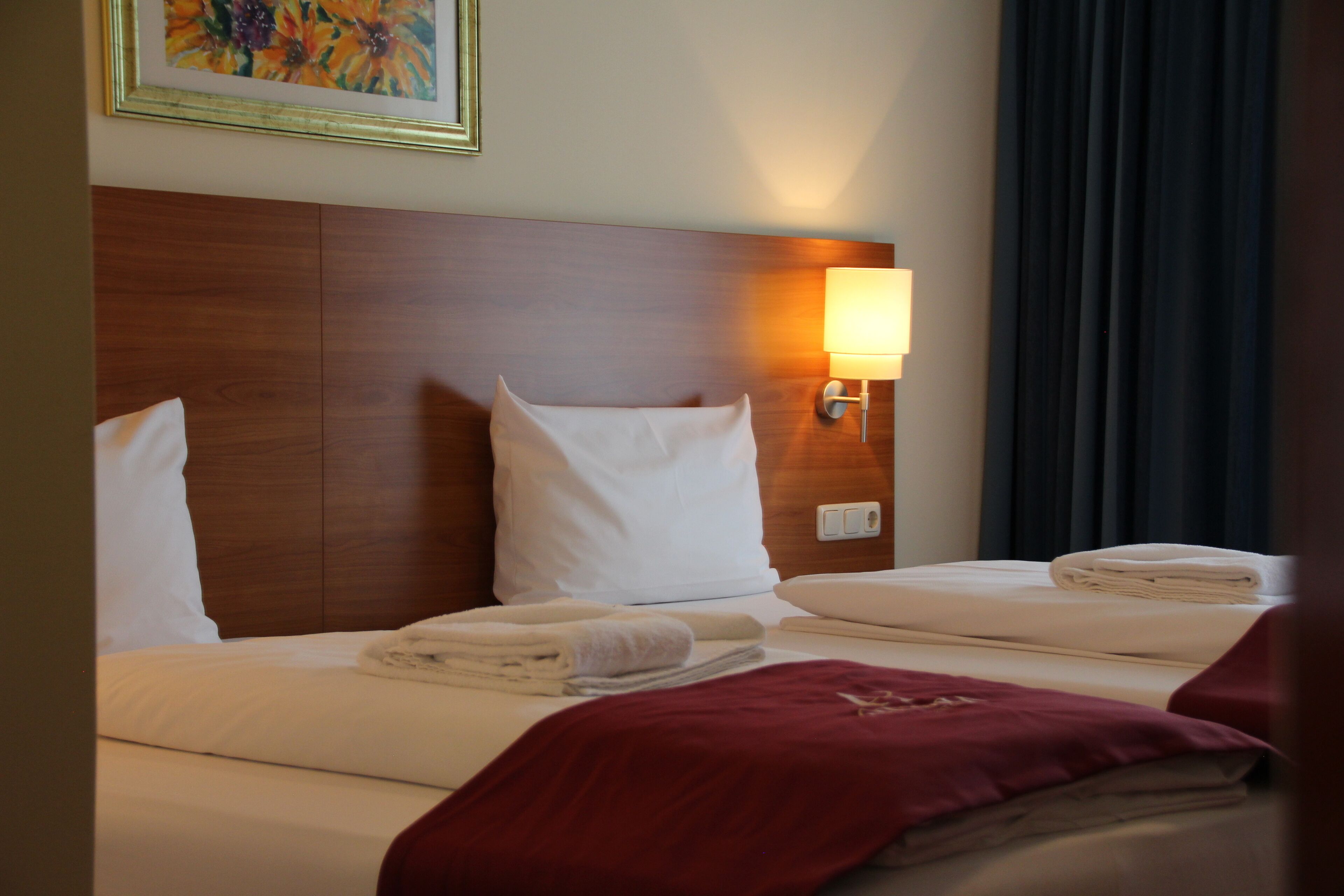 Une chambre d’hôtel avec une tête de lit en bois, deux lits avec du linge de lit blanc, un plaid rouge et une lampe fixée au mur.