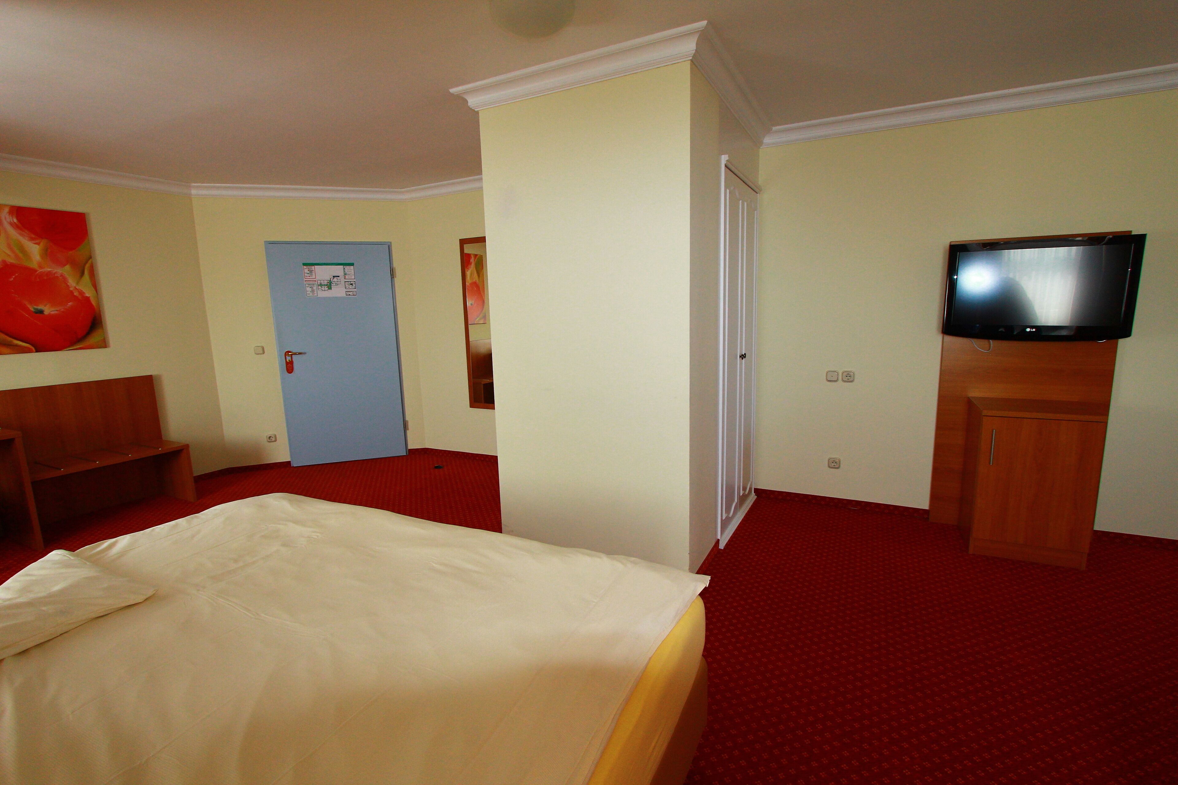 A hotel room with a bed, a television, a door, and a red carpeted floor.