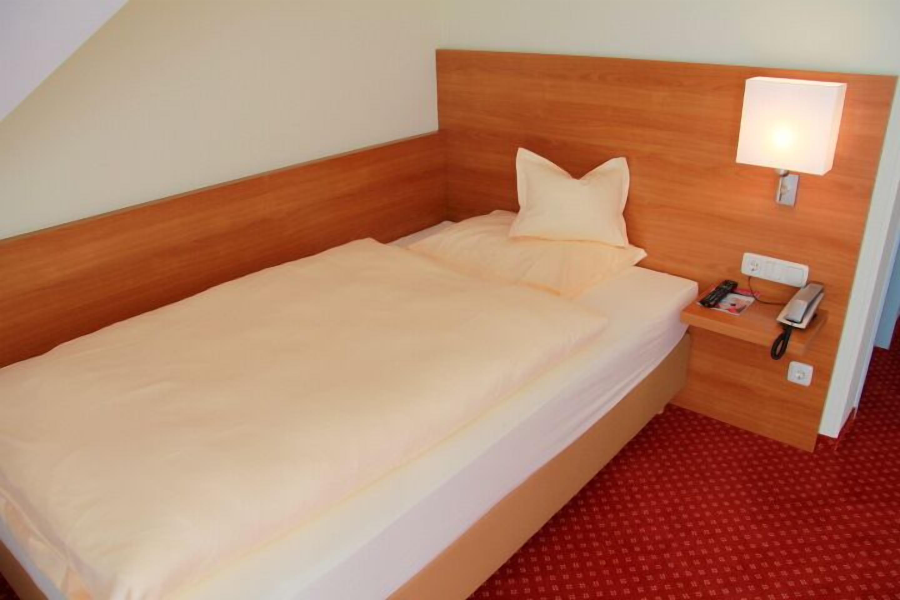 Una cama individual con cabecera de madera, una mesita de noche con lámpara y una alfombra roja estampada.