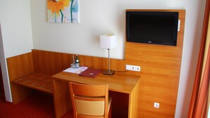 Hotellihuone, jossa on puinen työpöytä, tuoli, seinälle asennettu litteänäyttöinen televisio, lamppu ja kukkakuvamaalaus.