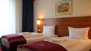 Hotellihuone, jossa on kaksi sänkyä, puinen pääty ja taulu seinällä sekä ikkuna, jossa on verhot.