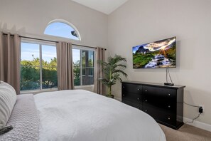 A bedroom with a large bed, a TV mounted on the wall, a potted plant, and a view of greenery through a window. - Luxury Private Townhome @ TPC | GOLF Garage & Pool (Scottsdale)