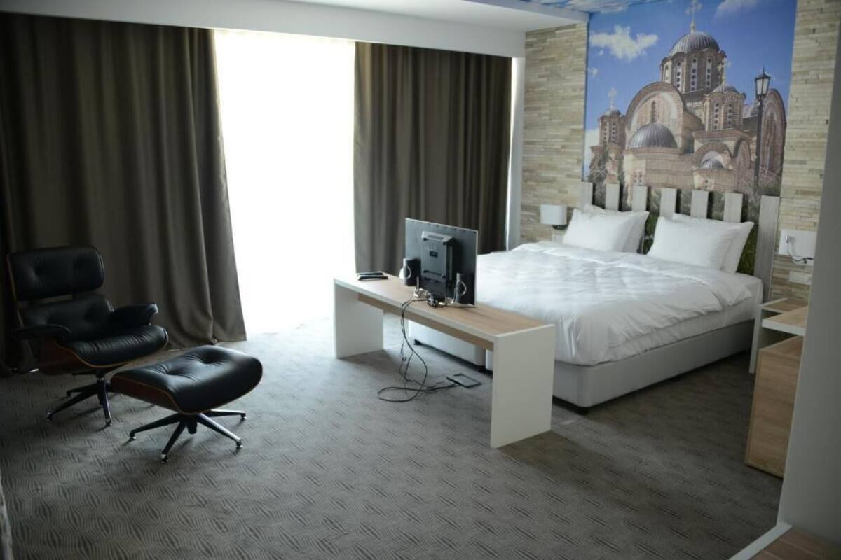 A hotel room with a bed, a desk with a computer, a chair, and a large window with curtains.