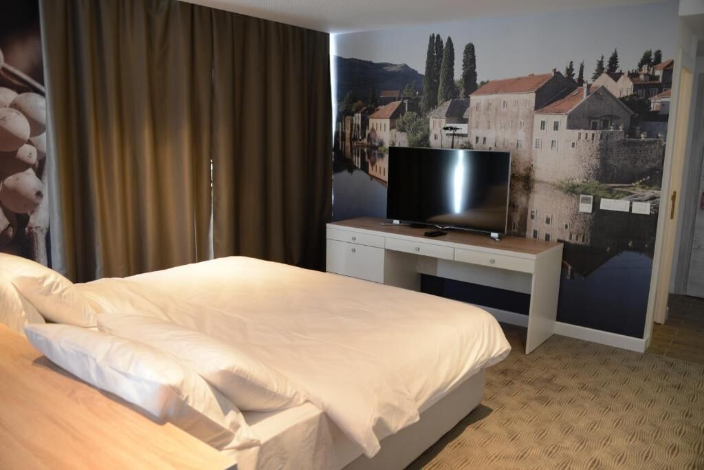 Une chambre d’hôtel avec un lit, un téléviseur à écran plat posé sur un bureau en bois, et une grande fresque murale représentant une scène de ville.