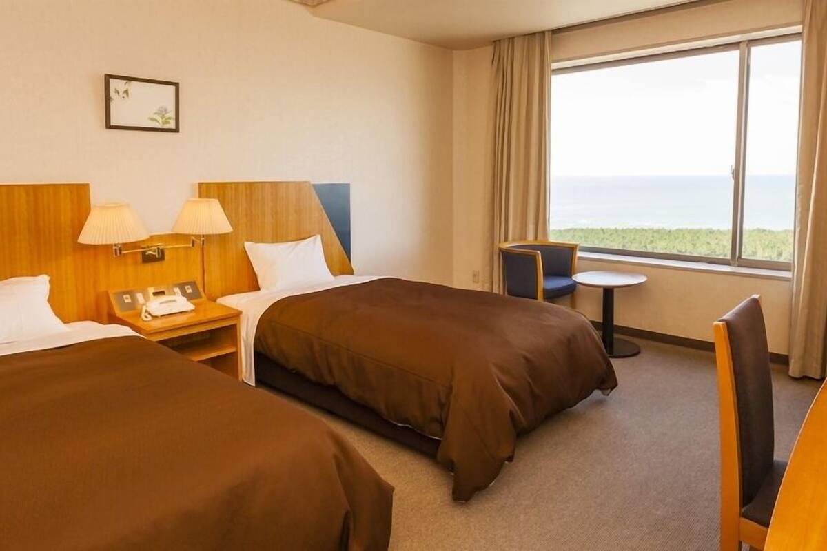Ett hotellrum med två sängar, ett skrivbord, en stol, ett litet bord och ett fönster med utsikt.