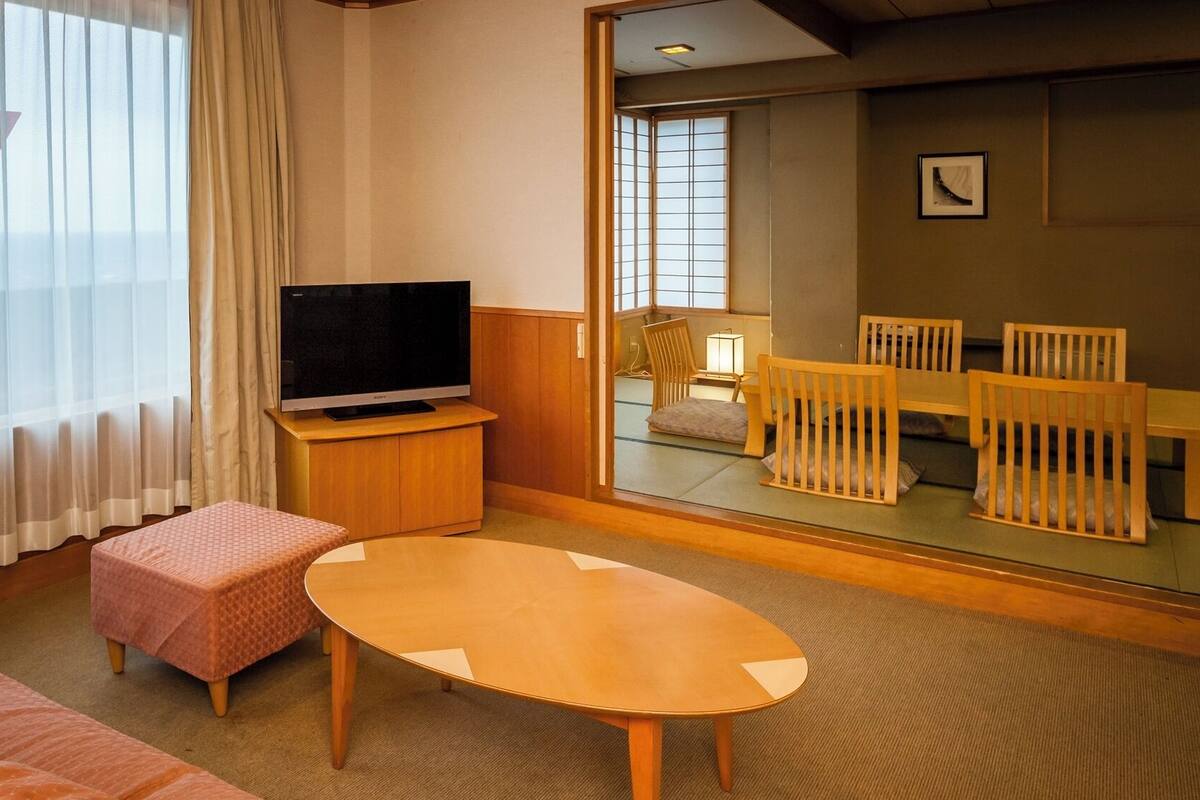 Ett hotellrum med en soffa, ett litet bord, en TV och utsikt över ett traditionellt japanskt rum med tatamimattor och ett lågt bord.