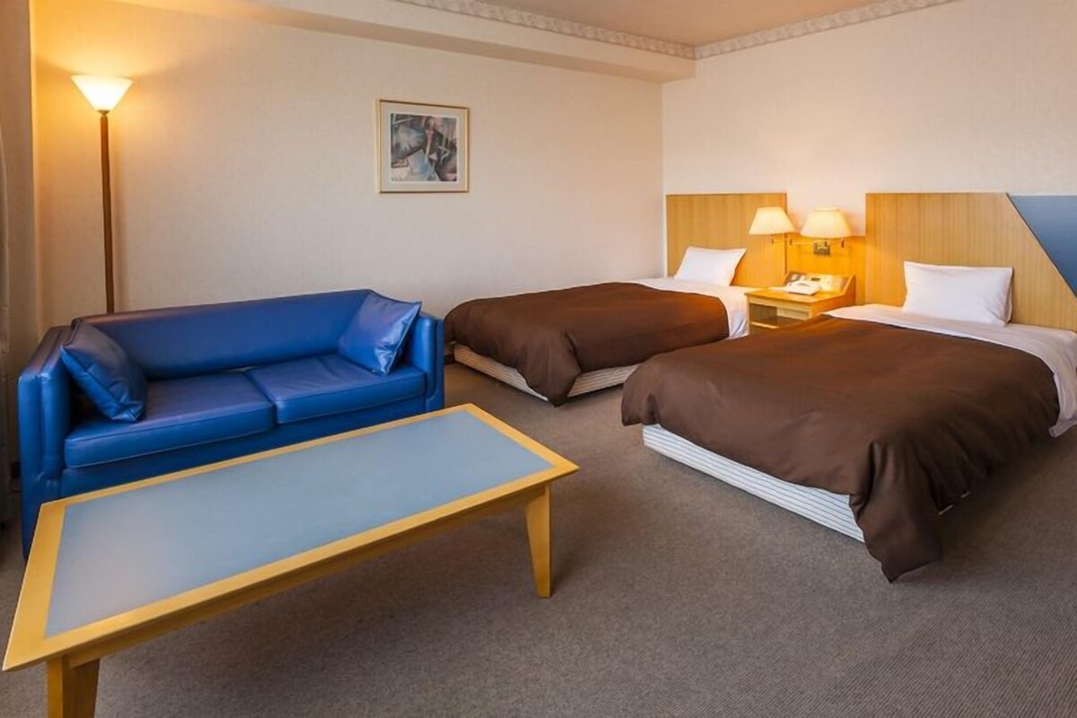 Habitación de hotel con un sofá azul, dos camas individuales, una mesita de noche de madera, una lámpara y un cuadro en la pared.