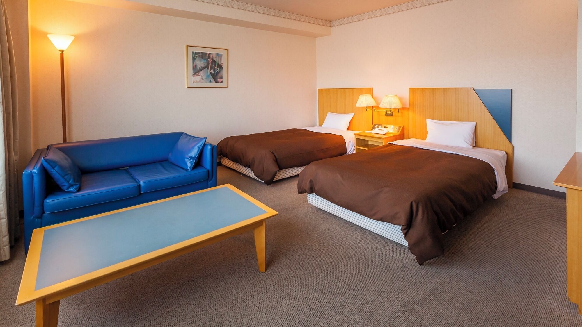 Et hotelværelse med en blå sofa, to enkeltsenge med brunt sengetøj, et trænatbord, et bord og en lampe.