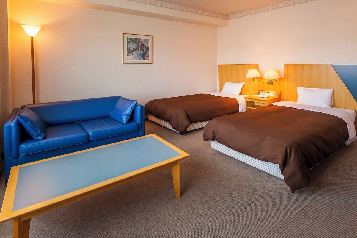 Ett hotellrum med en blå soffa, två enkelsängar med bruna sängkläder, ett träbord som sängbord, ett bord och en lampa.