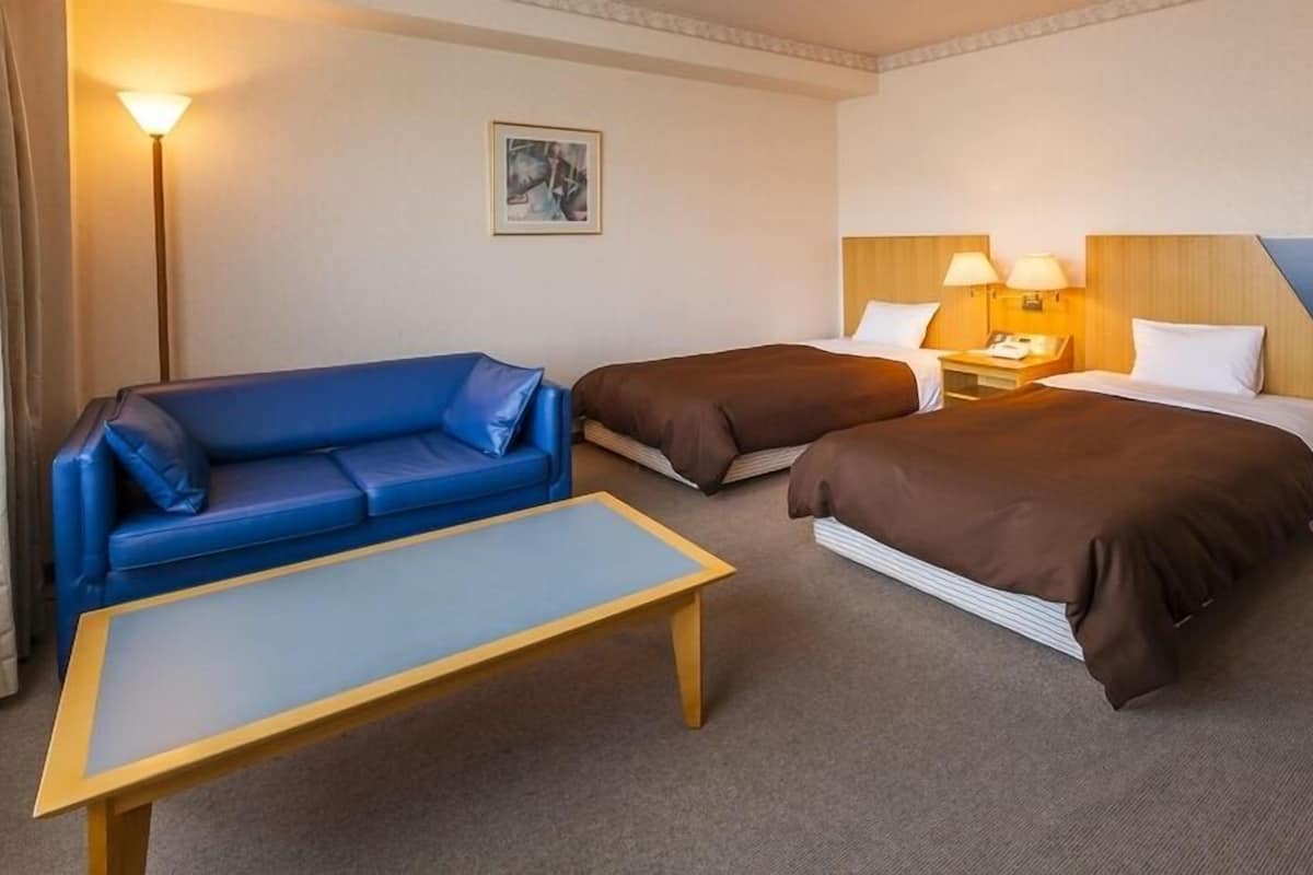 Ett hotellrum med en blå soffa, två enkelsängar, ett träbord vid sängen, en lampa och en tavla på väggen.