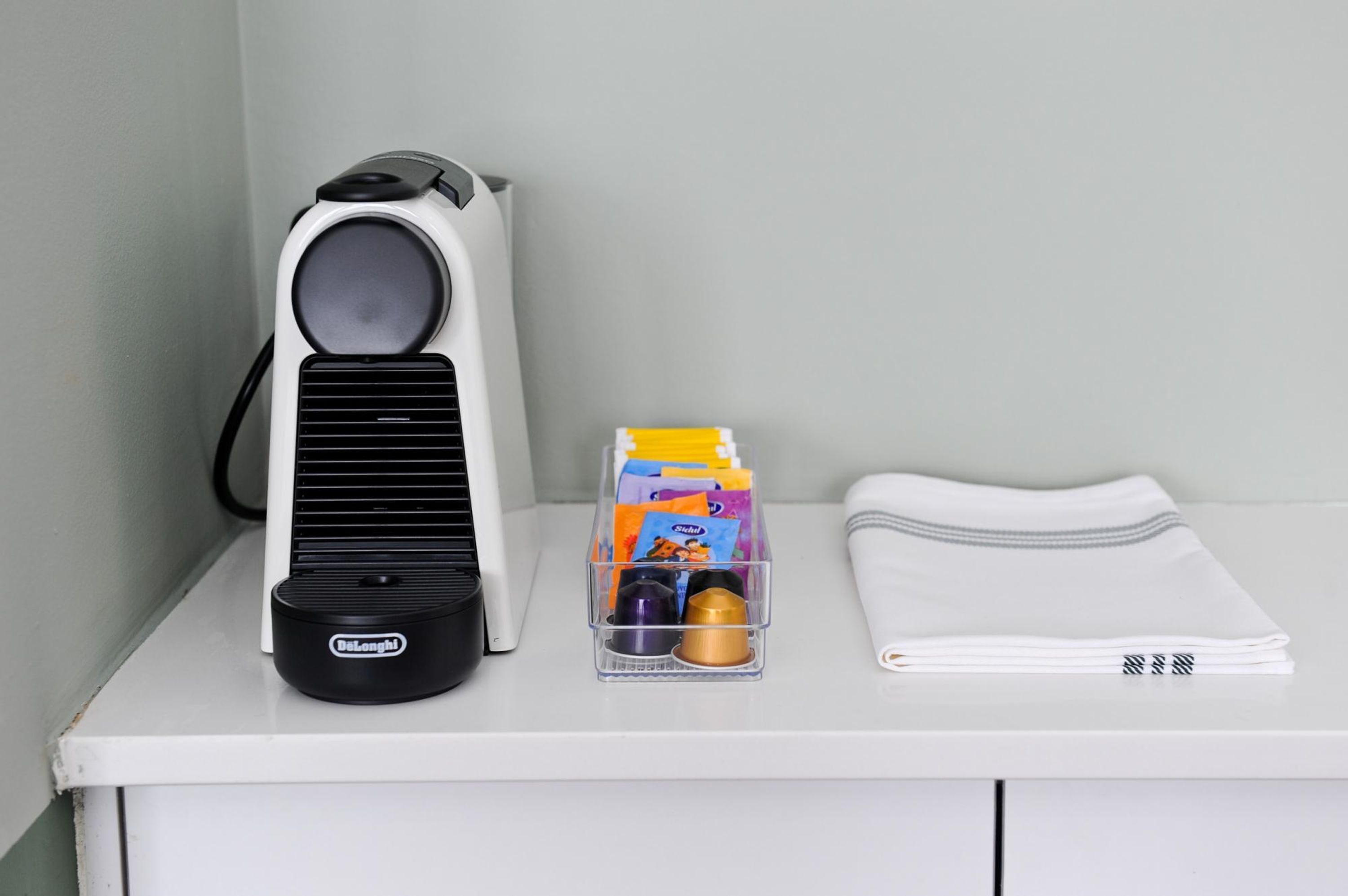 Un bureau blanc avec une machine à café DeLonghi, un organiseur transparent en plastique contenant du matériel de bureau et une serviette blanche soigneusement pliée.