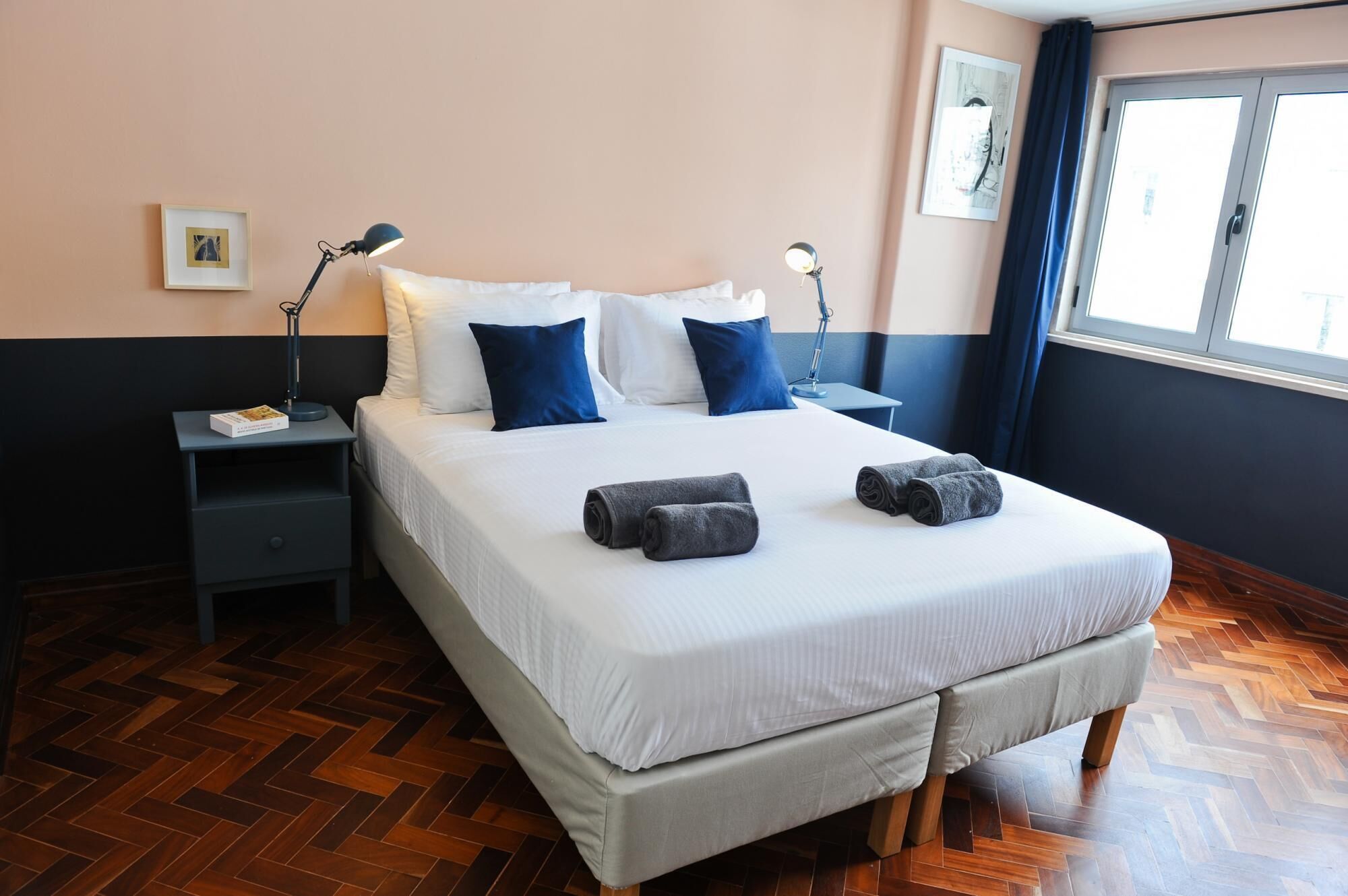 A neatly made bed with white and blue pillows, two bedside lamps, a small bedside table with a book, and a window with curtains.