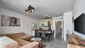 Un salon moderne avec un grand canapé d’angle beige, un coin repas avec une table et des chaises en bois, et une cuisine en arrière-plan.