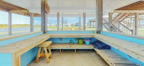 A covered outdoor seating area with wooden benches, a small table, and a view of the beach and houses.