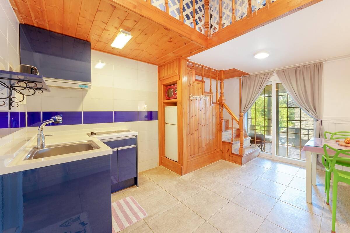 A modern kitchen with a wooden staircase, a dining area with green chairs, and large windows.